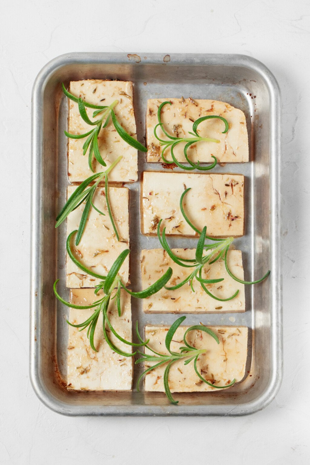 An overhead image of a small, silver baking dish, which has been filled with tofu slices and fresh rosemary prior to baking.