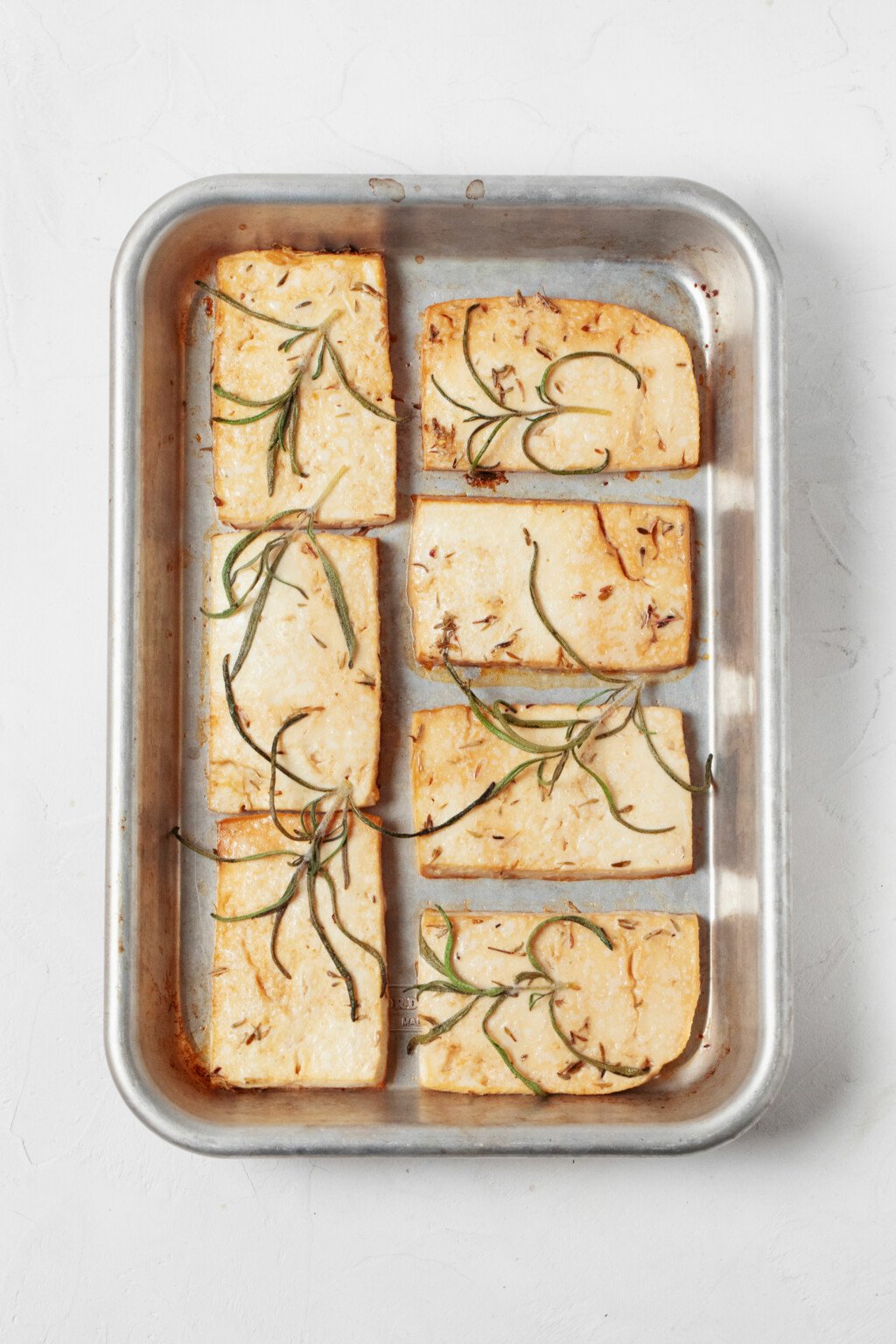 An overhead image of baked tofu slices, covered with long, thin pieces of fresh rosemary.