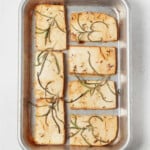 An overhead image of a small, silver baking sheet, which has been lined with tofu slices that are topped with green rosemary leaves.