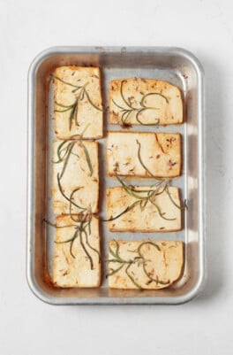 An overhead image of a small, silver baking sheet, which has been lined with tofu slices that are topped with green rosemary leaves.