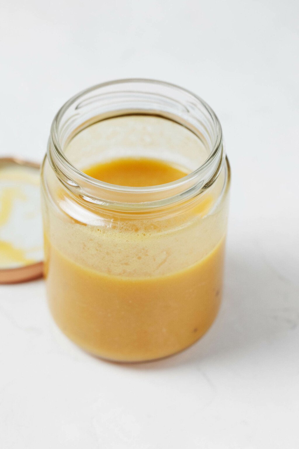 A glass mason jar rests on a white surface. It is filled with a creamy, pale golden vinaigrette.