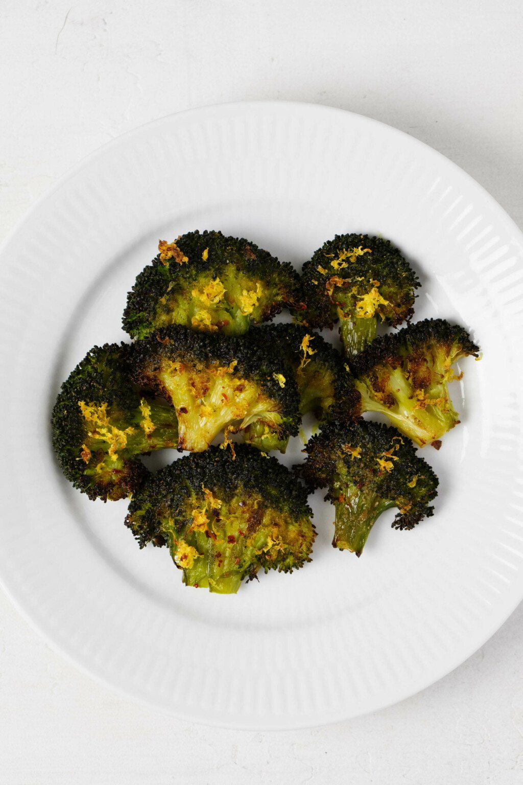 Broccoli florets have been flattened and roasted till crisp tender, then topped with lemon zest. They rest on a white plate.
