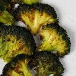 Broccoli florets have been flattened and roasted till crisp tender, then topped with red chili flakes. They rest on a white plate.