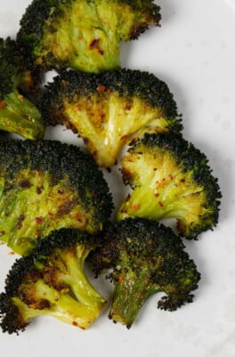 Broccoli florets have been flattened and roasted till crisp tender, then topped with red chili flakes. They rest on a white plate.