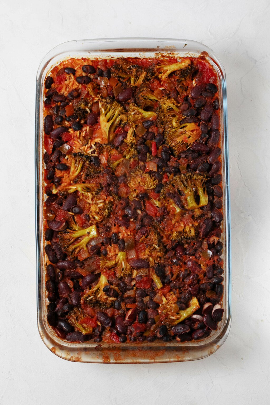 An overhead image of a baked rice and bean dish. The top is has darkened tomatoes and little pieces of broccoli visible.