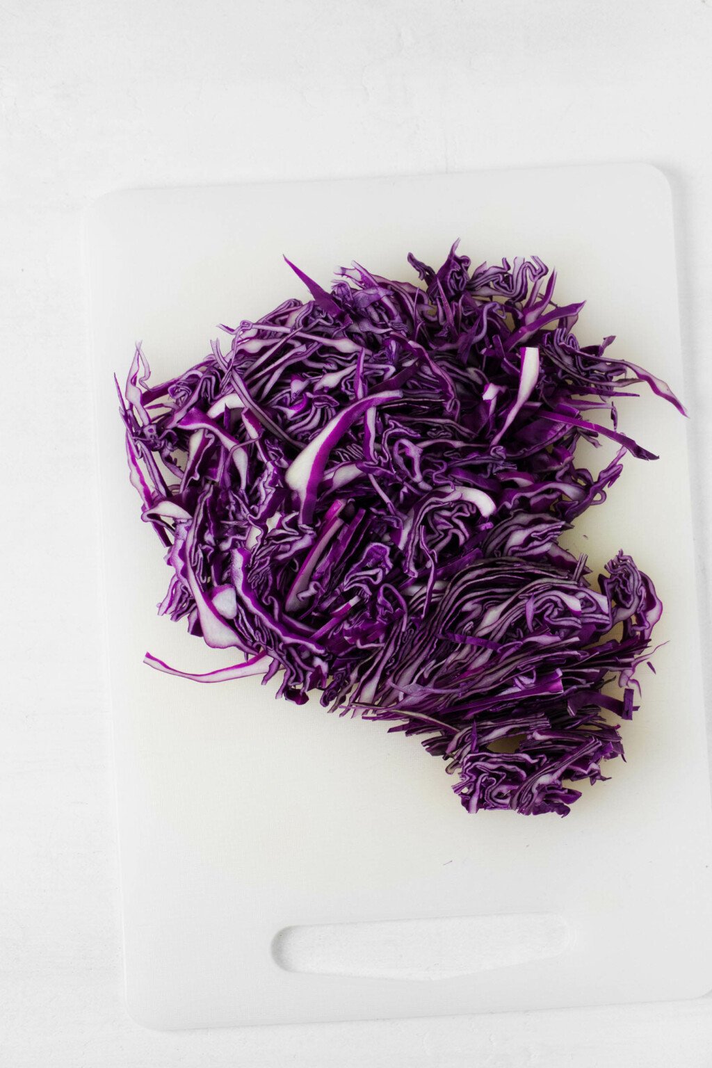Shredded red cabbage is pictured on a small, white cutting board. 