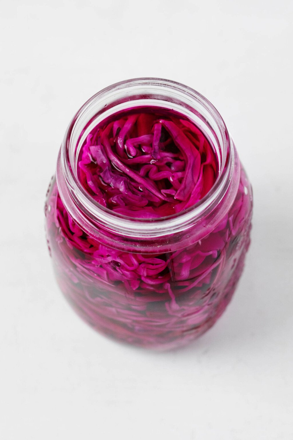 An image of a large, white mason jar, which has been filled with shredded red cabbage in a brine. It rests on a white surface.