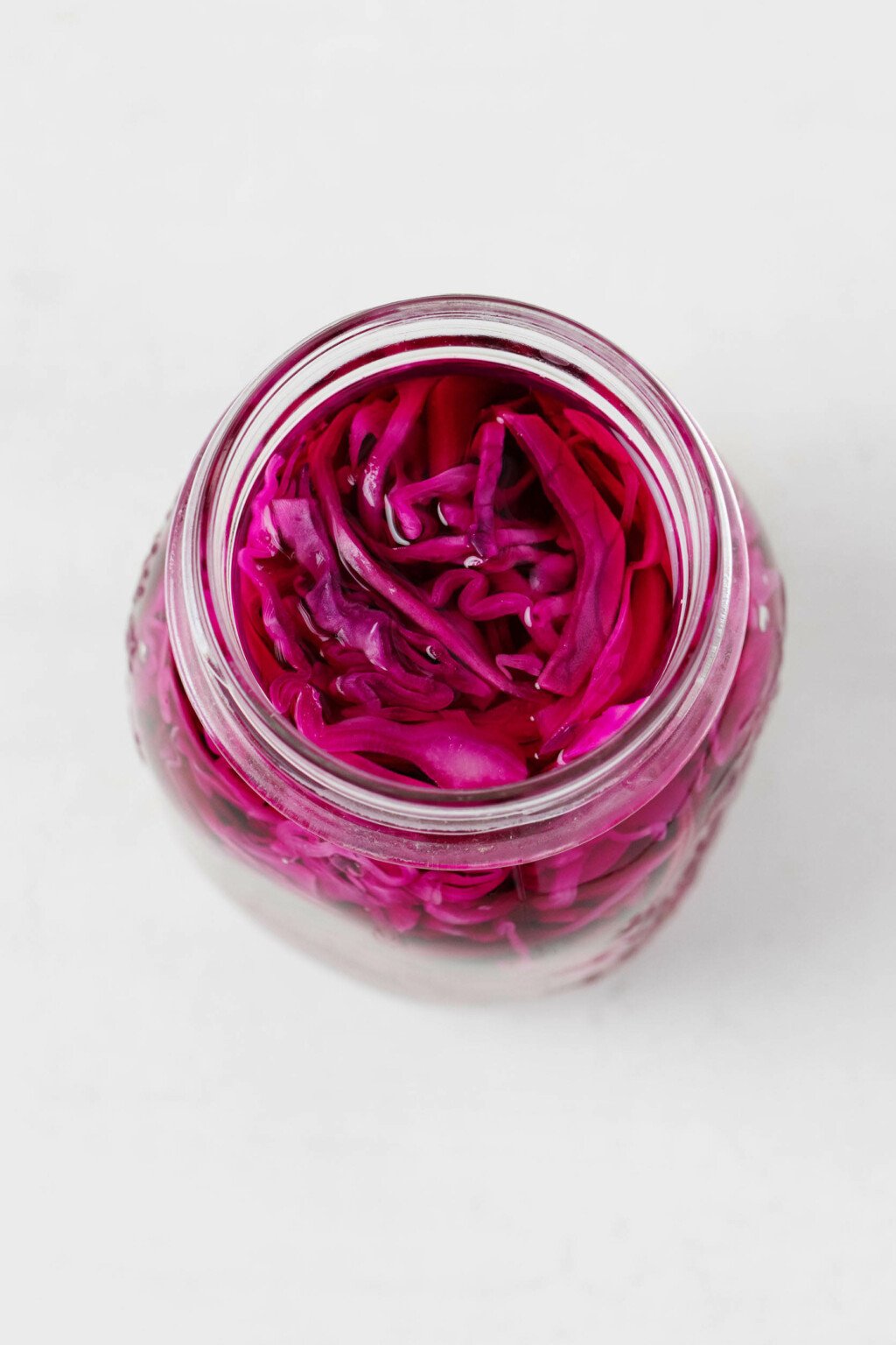 An image of a large, white mason jar, which has been filled with shredded red cabbage in a brine. It rests on a white surface.