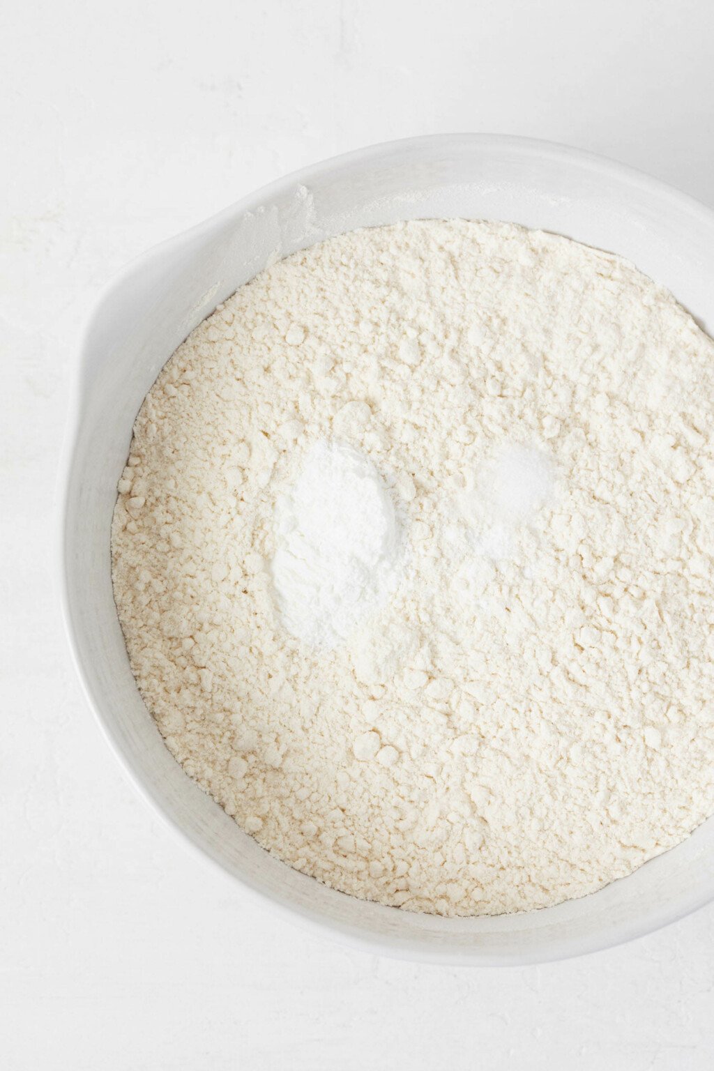 Flour and sugar are pictured overhead in a large, white mixing bowl. 