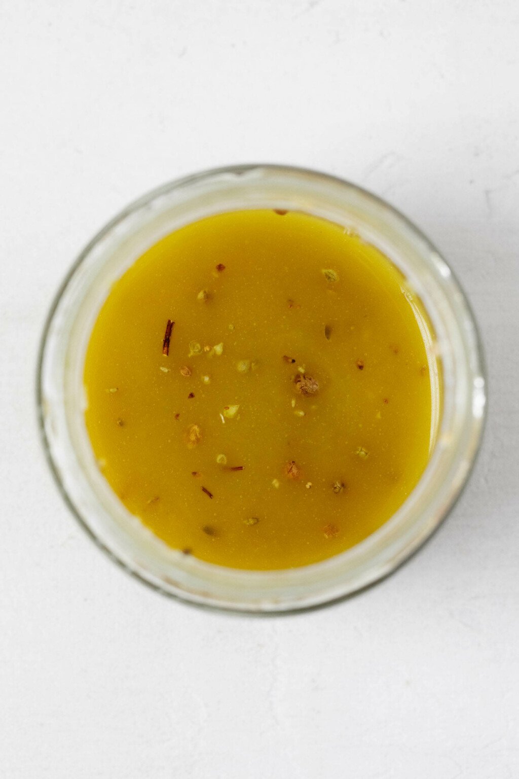 An overhead image of a glass jar, which contains a vinaigrette that is flecked with dried herbs.