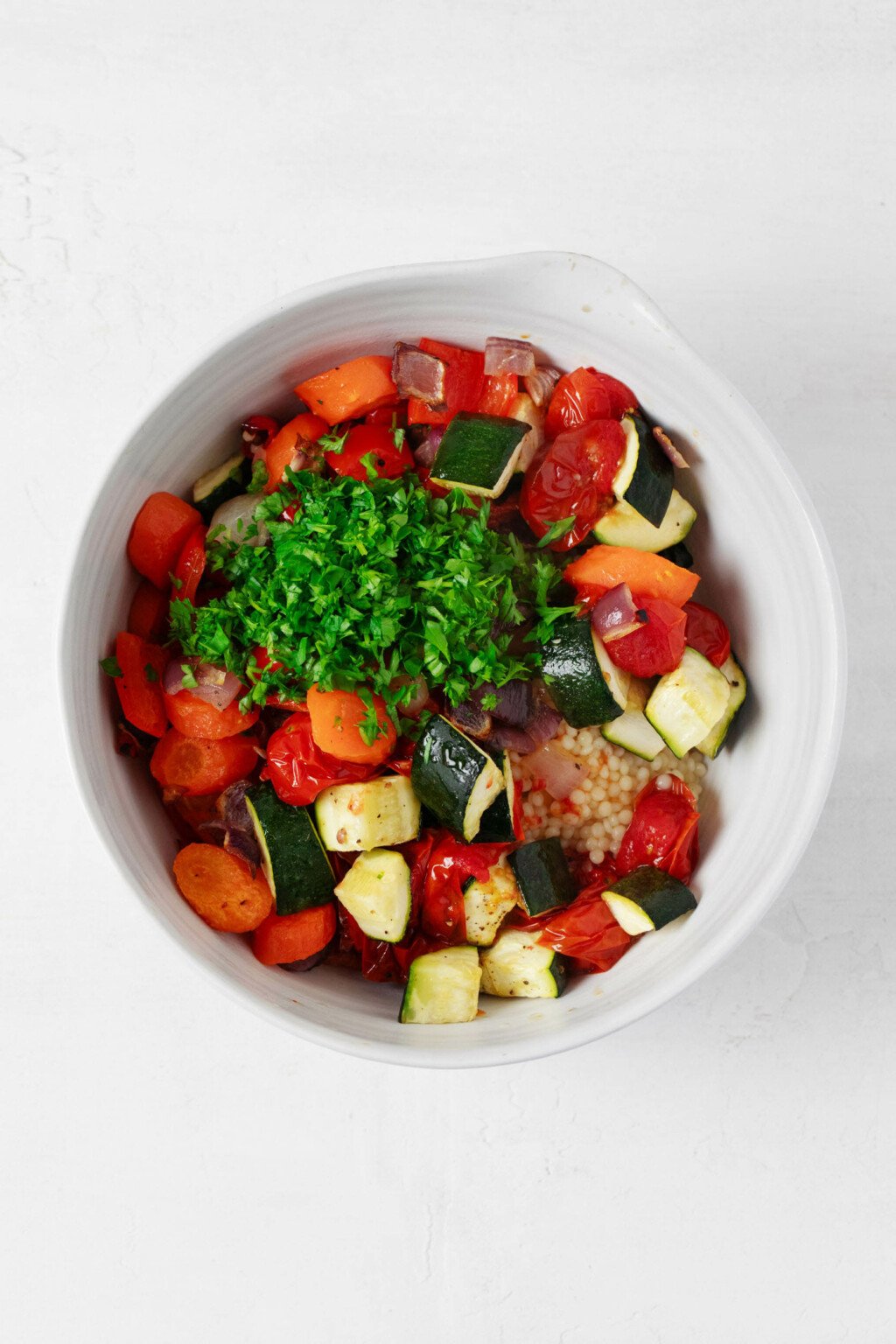 An overhead image of a mixing bowl that contains a bed of couscous and a medley of roasted vegetables, as well as bright green, chopped parsley.