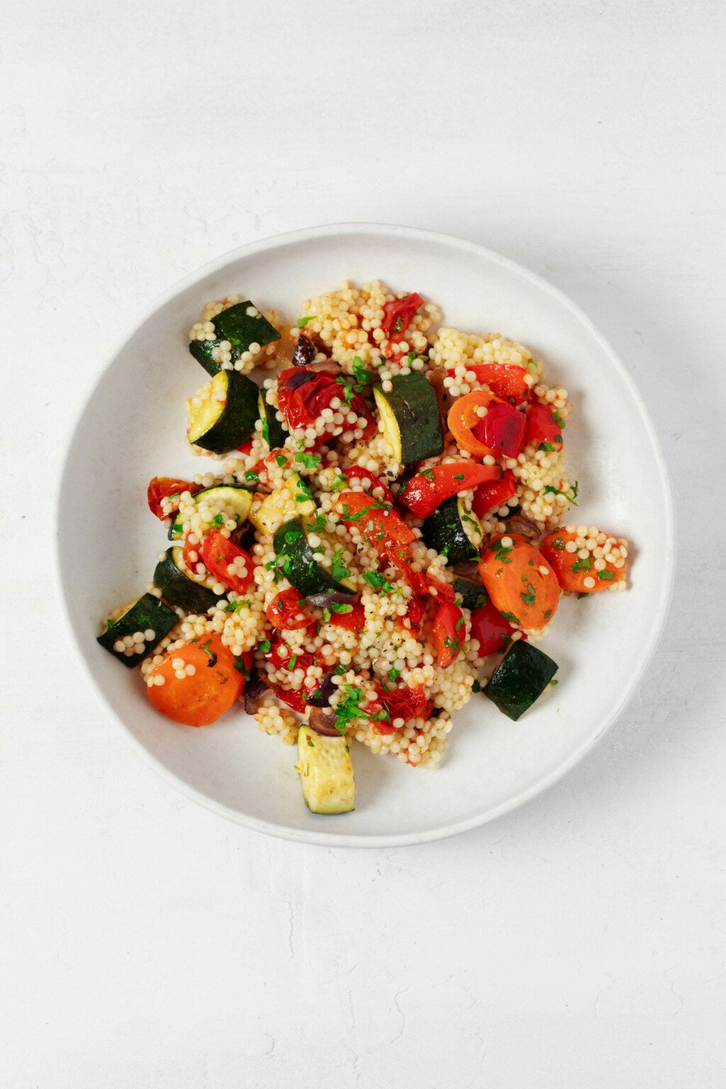 A roasted vegetable and pearl couscous salad has been arranged in a shallow, round white bowl. It rests on a white surface.