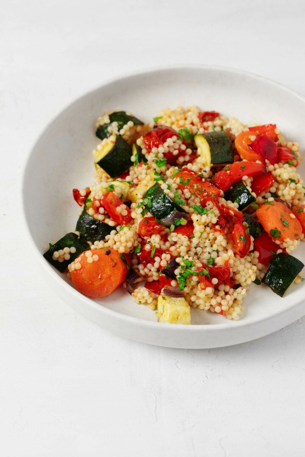 A roasted vegetable and pearl couscous salad has been arranged in a shallow, round white bowl. It rests on a white surface.