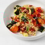 A roasted vegetable and pearl couscous salad has been arranged in a shallow, round white bowl. It rests on a white surface.