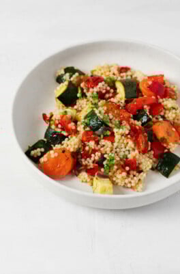 A roasted vegetable and pearl couscous salad has been arranged in a shallow, round white bowl. It rests on a white surface.