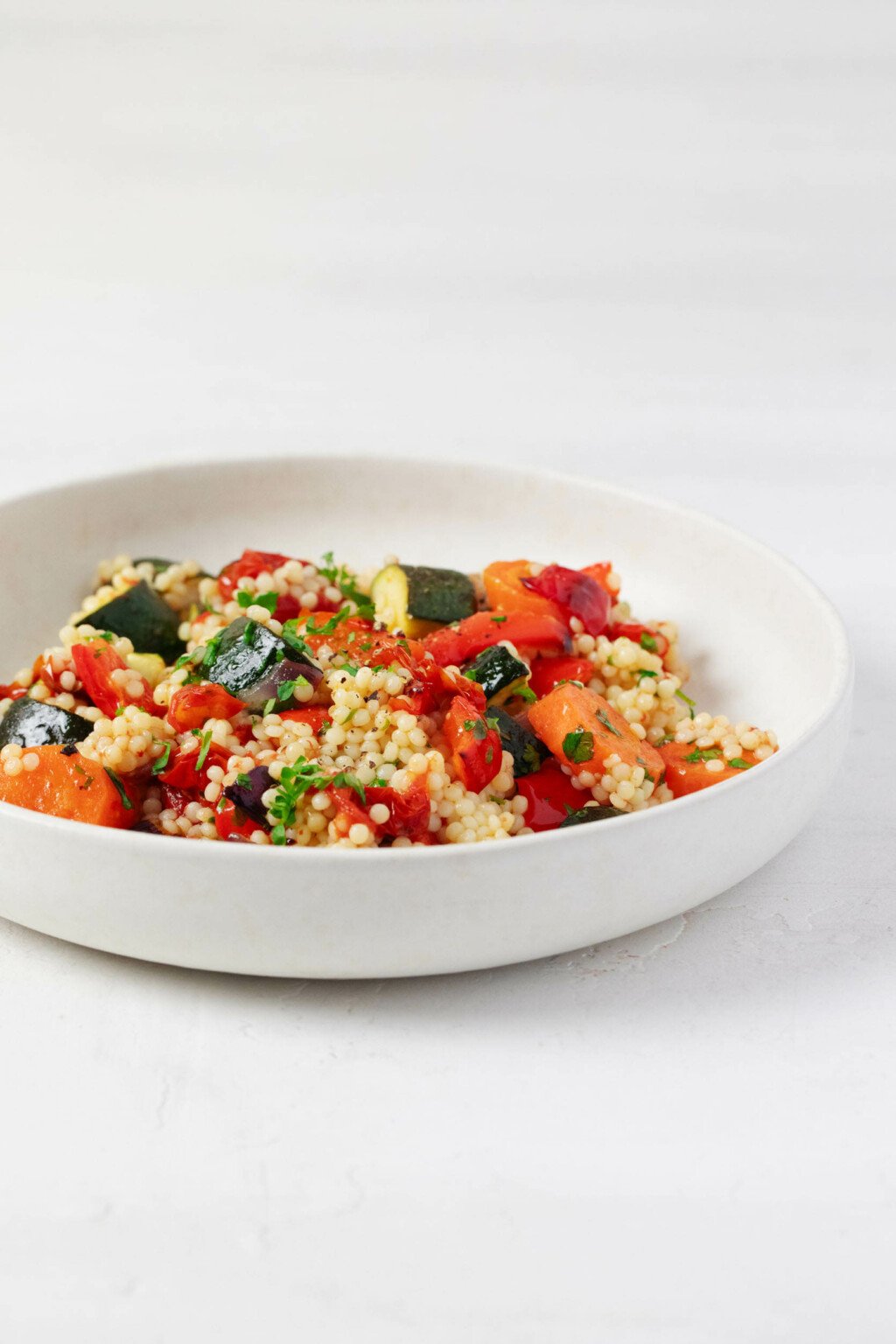 A roasted vegetable and pearl couscous salad has been arranged in a shallow, round white bowl. It rests on a white surface.