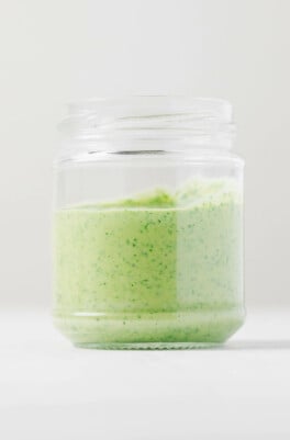 An image of a glass jar resting on a white surface. The jar has been filled with creamy tahini pesto.