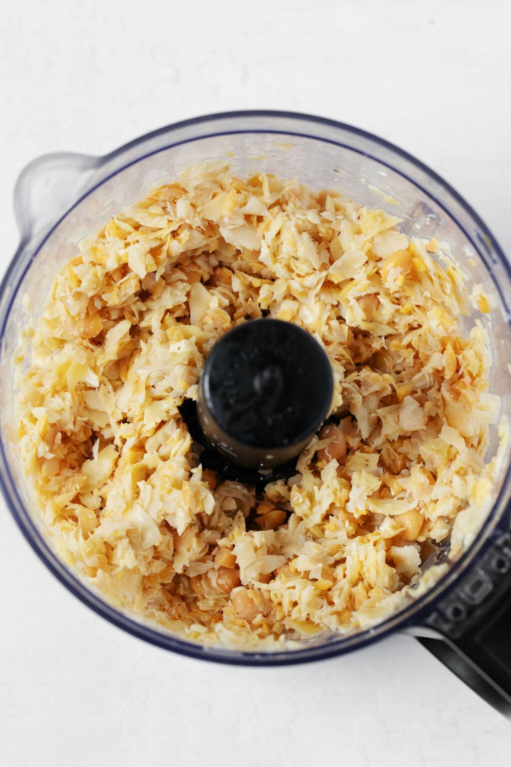 An overhead image of the bowl of a food processor, which has been fitted with the "S" blade. The processor contains finely chopped up chickpeas.