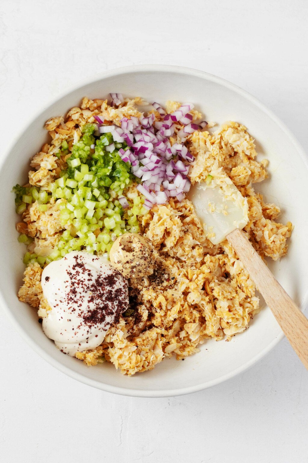 A white mixing bowl has been filled with chopped artichoke hearts, chickpeas, diced red onion, celery, mayonnaise, and seaweed flakes.