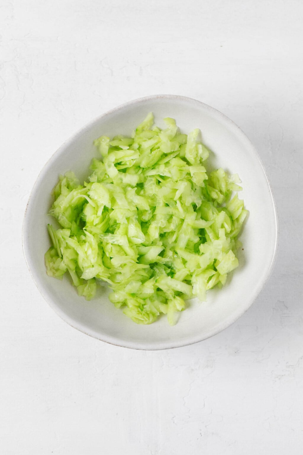 An asymmetrical white bowl is filled with grated cucumber.