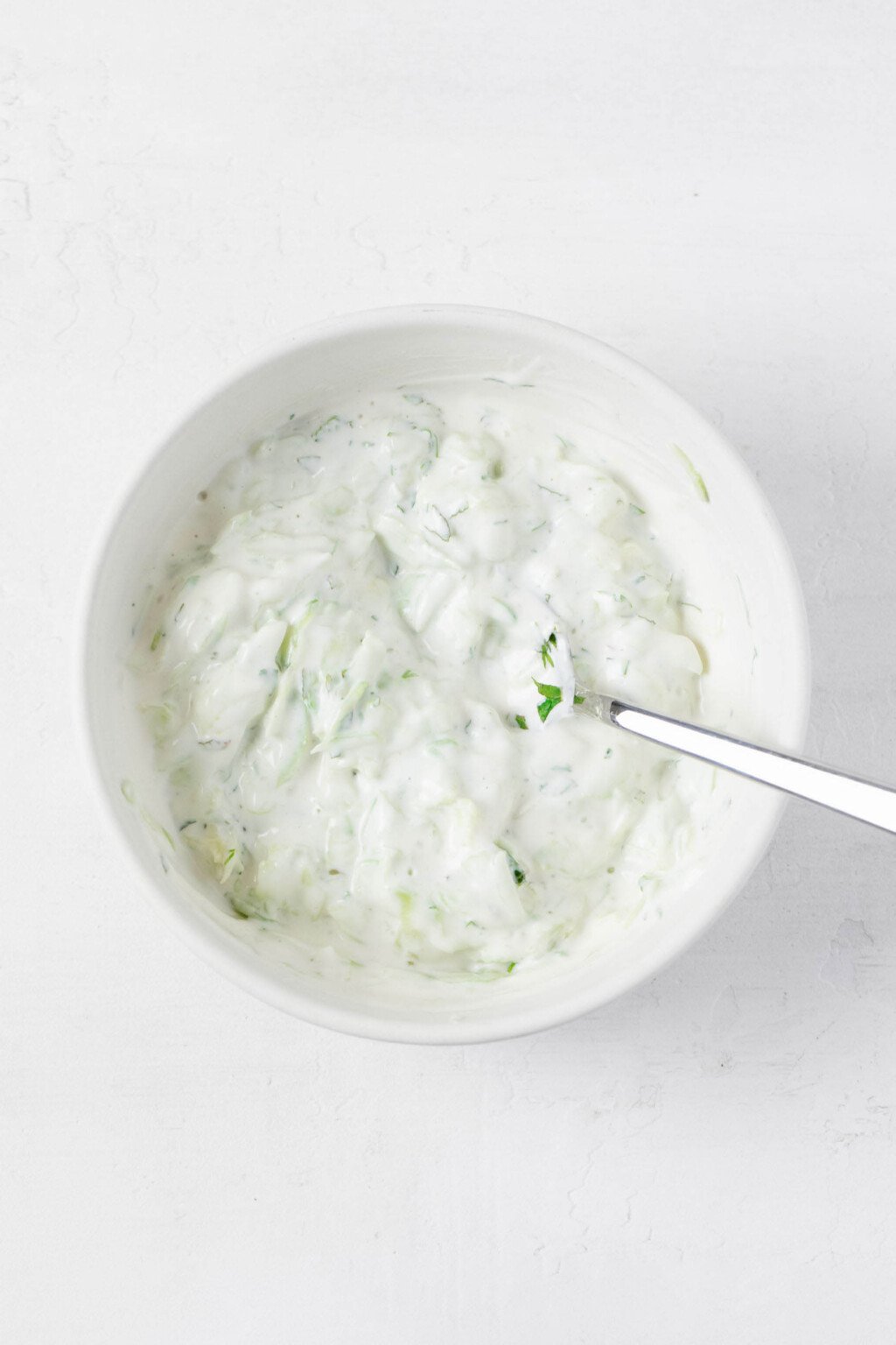 An overhead image of a round, white bowl, which is filled with a creamy vegan verson of tzatziki dip.