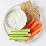 An image of a round, white bowl of vegan tzatziki dip, which is served with raw vegetables and crackers.