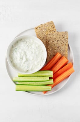 An image of a round, white bowl of vegan tzatziki dip, which is served with raw vegetables and crackers.