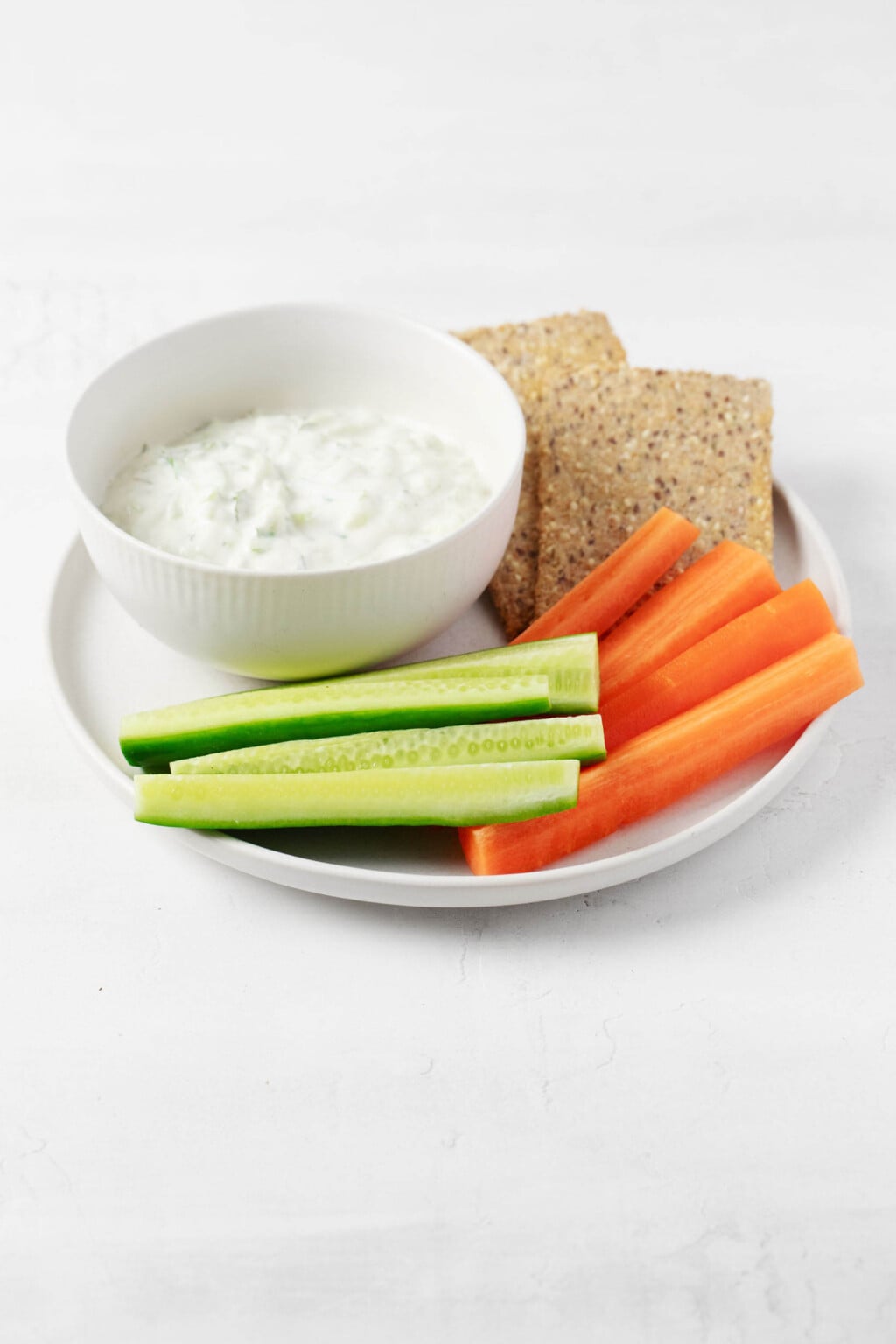 An image of a round, white bowl of vegan tzatziki dip, which is served with raw vegetables and crackers.