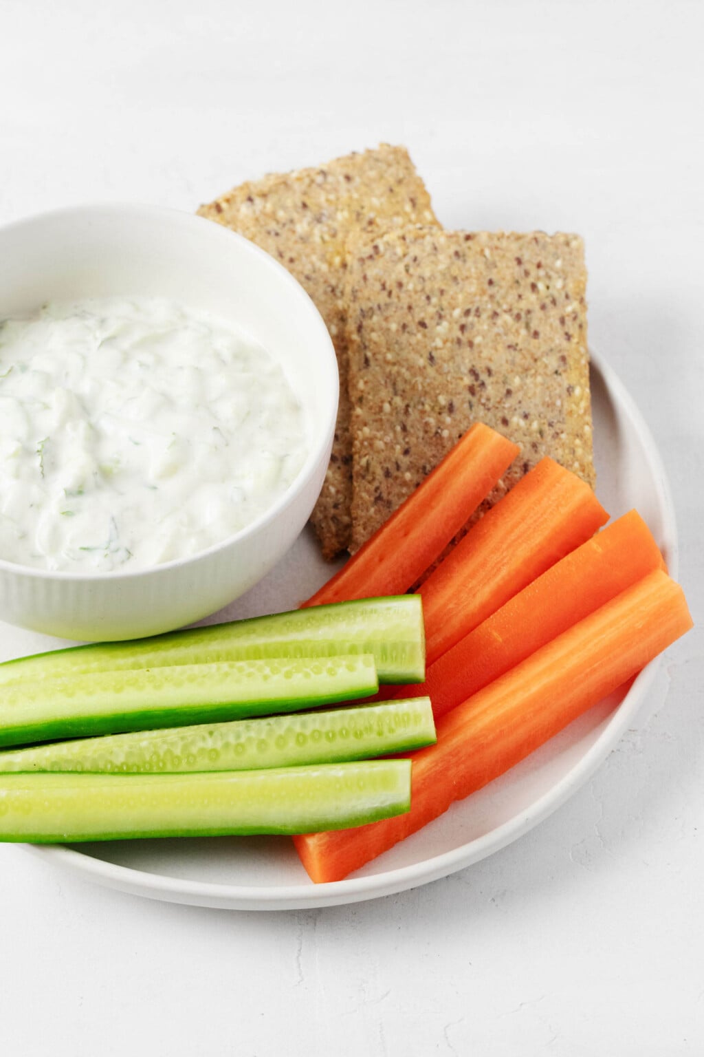 An image of a round, white bowl of vegan tzatziki dip, which is served with raw vegetables and crackers.