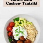 A vibrant vegan Greek bowl is being assembled in a white ceramic vessel. The bowl includes orzo, fresh vegetables, olives, and chickpea oat balls.
