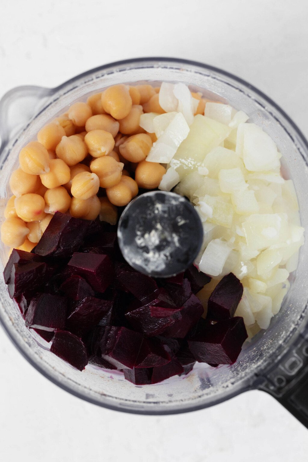 Chickpeas, onion, and cooked beets have been added to the clear bowl of a food processor.