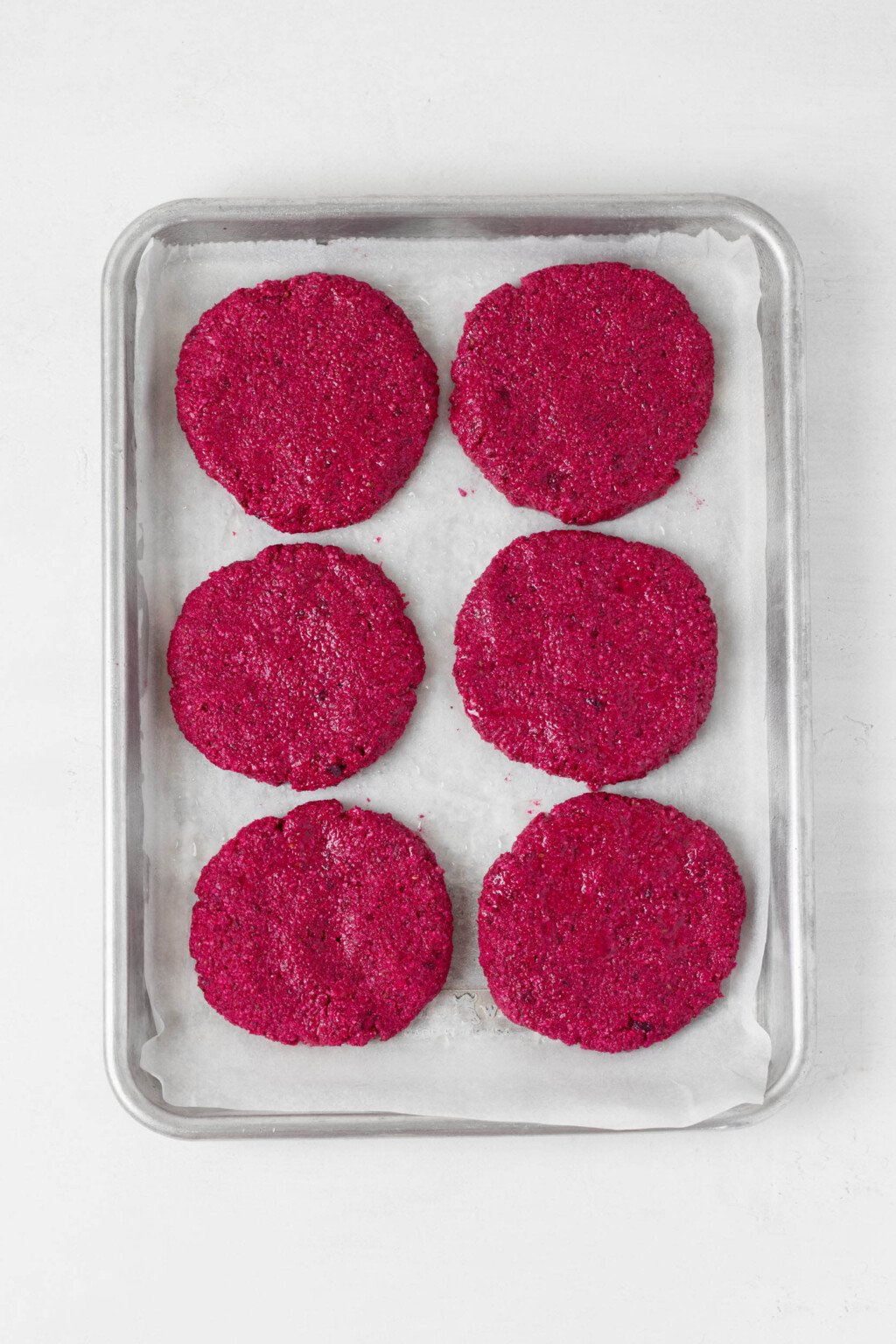 Six bright red beet burger patties have been arranged onto a parchment-lined baking sheet.