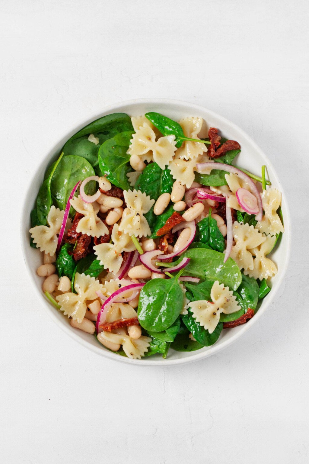 A round, white ceramic shallow bowl has been filled with a fresh spinach pasta salad, made with fresh green leaves of baby spinach and red sun-dried tomatoes.