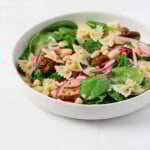 A round, white ceramic shallow bowl has been filled with a fresh spinach pasta salad, made with fresh green leaves of baby spinach and red sun-dried tomatoes.