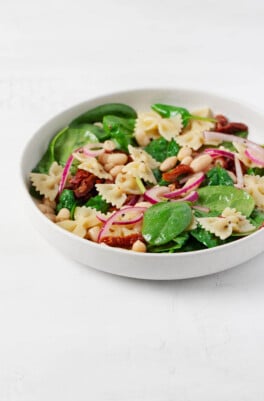 A round, white ceramic shallow bowl has been filled with a fresh spinach pasta salad, made with fresh green leaves of baby spinach and red sun-dried tomatoes.