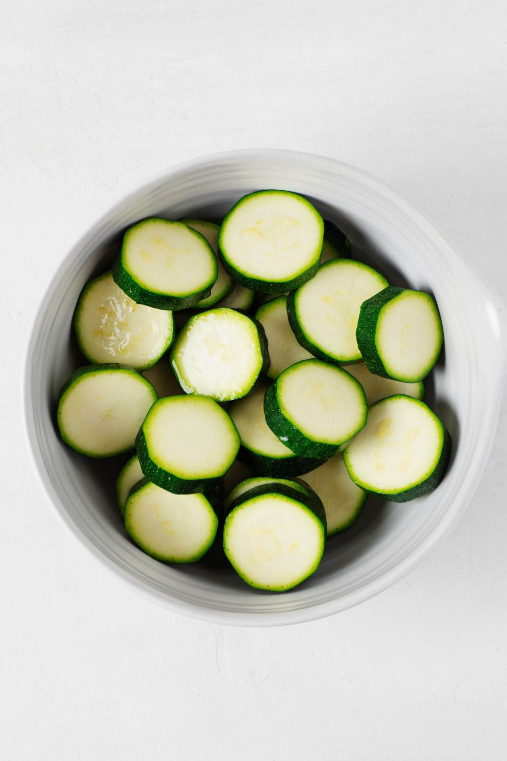 A raw zucchini has been sliced into thin, round pieces, which are arranged in a white bowl.