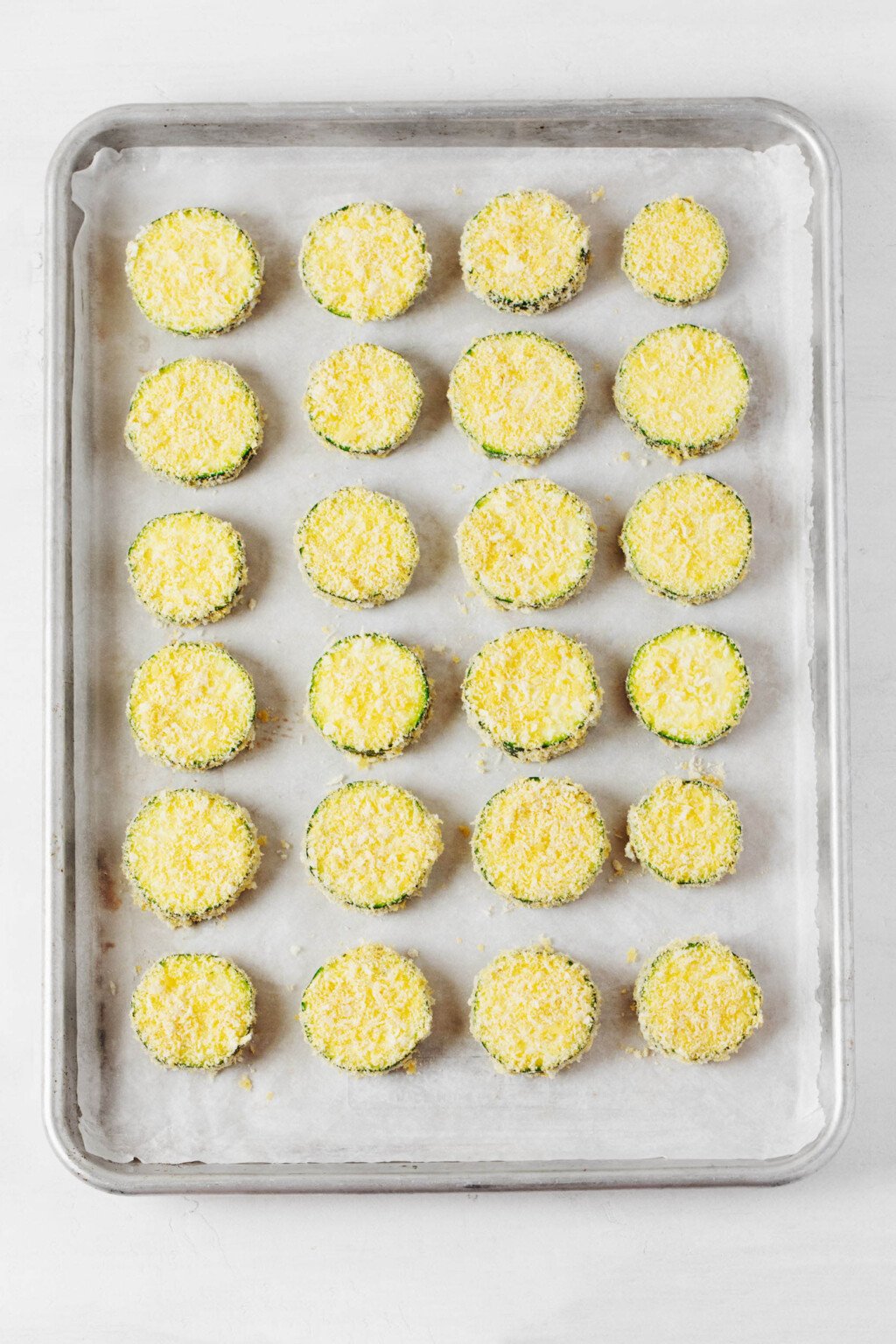 Round pieces of zucchini, coated with bread crumbs and almond flour, have been arranged on a baking sheet in preparation for baking.