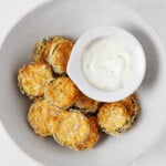 Oven-baked, lightly browning, round vegan zucchini chips are piled into a round, white bowl, along with a white-colored dipping sauce.