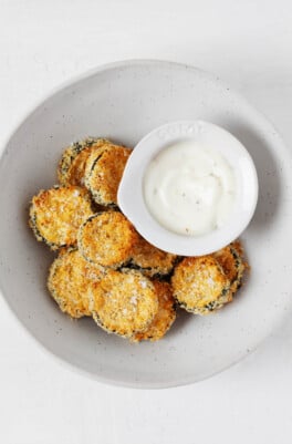Oven-baked, lightly browning, round vegan zucchini chips are piled into a round, white bowl, along with a white-colored dipping sauce.