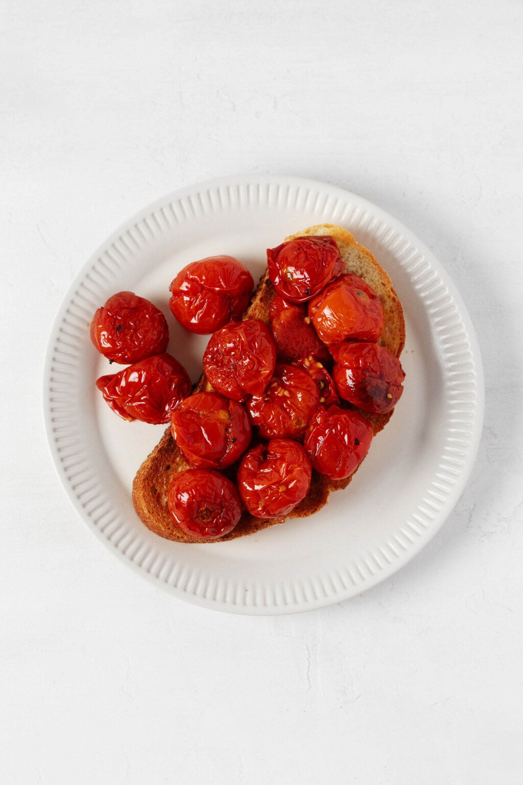 A slice of roasted bread has been piled high with red, oven-roasted cherry tomatoes. The bread is arranged on a round, fluted white plate.