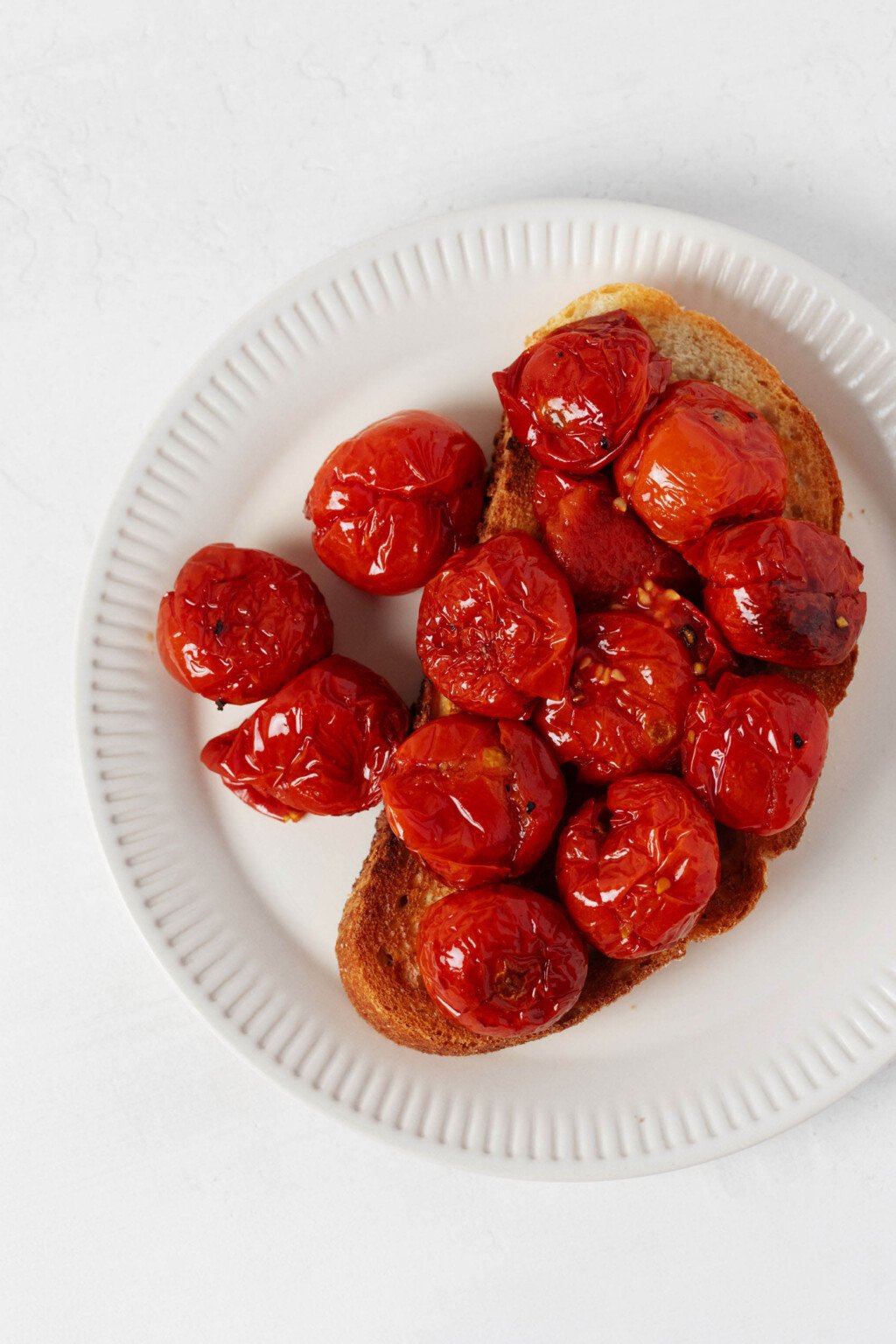 A slice of roasted bread has been piled high with red, oven-roasted cherry tomatoes. The bread is arranged on a round, fluted white plate.
