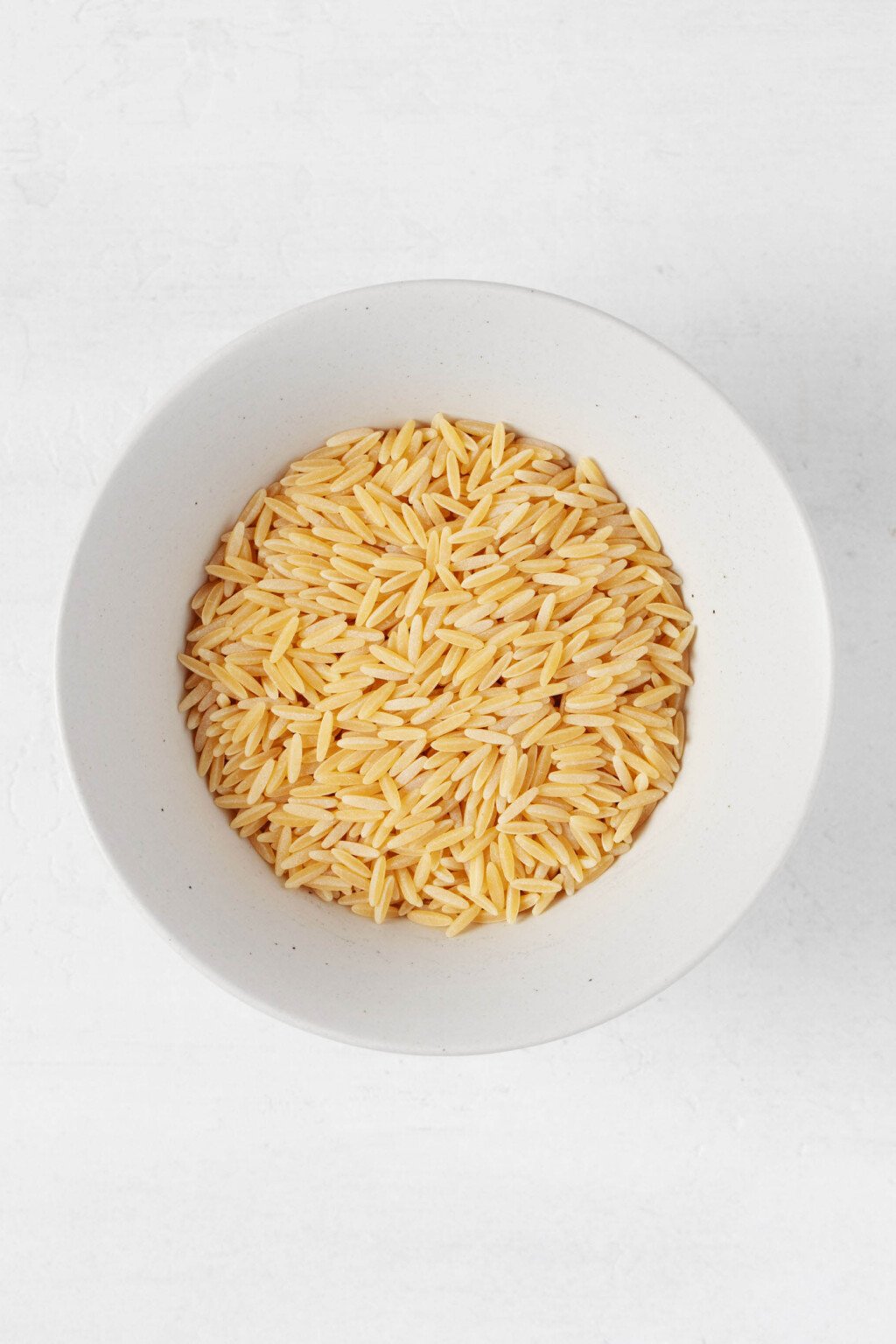 An overhead image of a white bowl, which is filled with uncooked orzo.