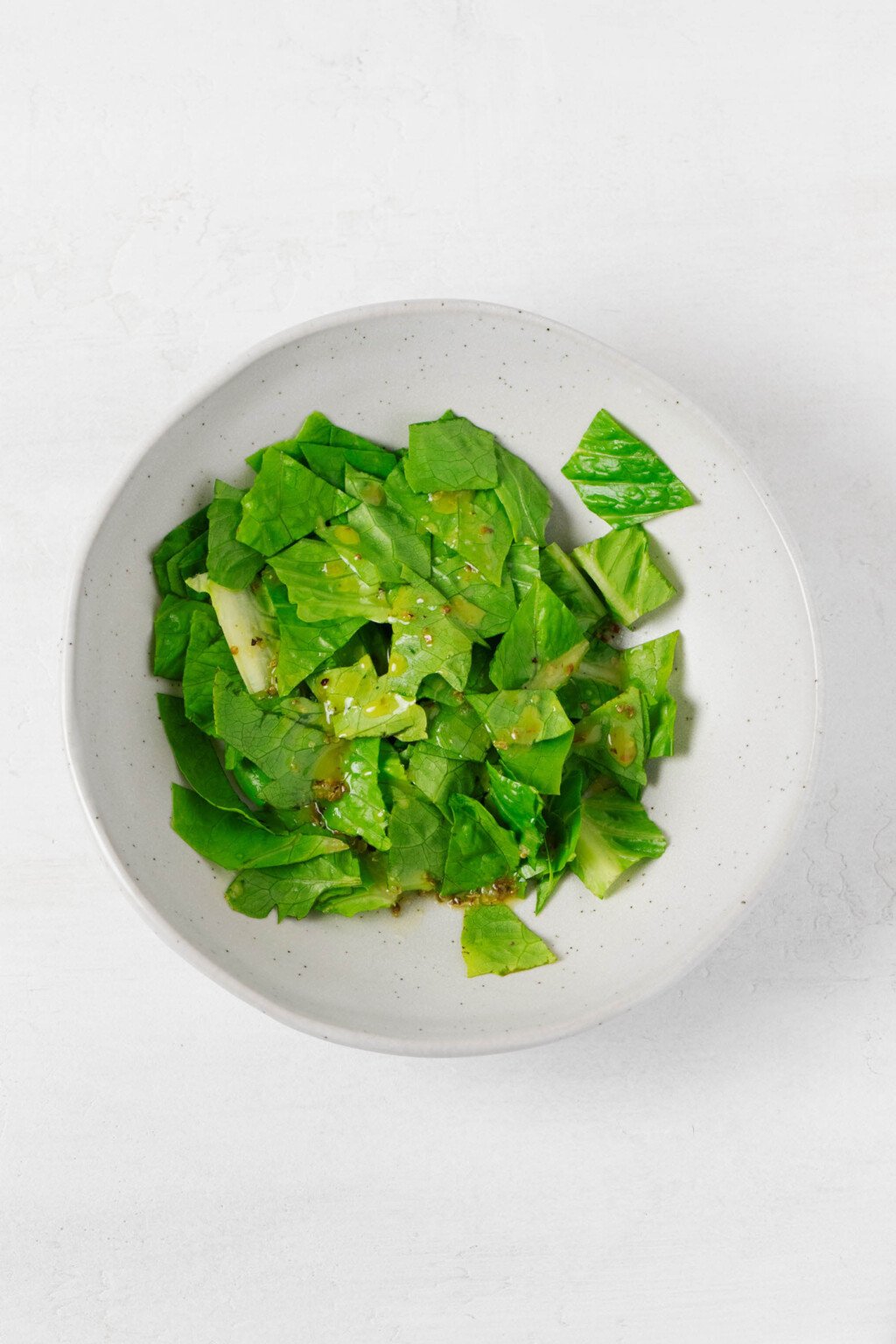 A round, white ceramic bowl is filled with greens that have been lightly dressed with vinaigrette.