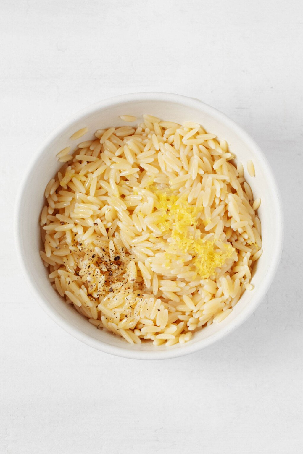 Finely grated lemon zest has been added to a bowl of cooked orzo, which is piled into a white bowl.