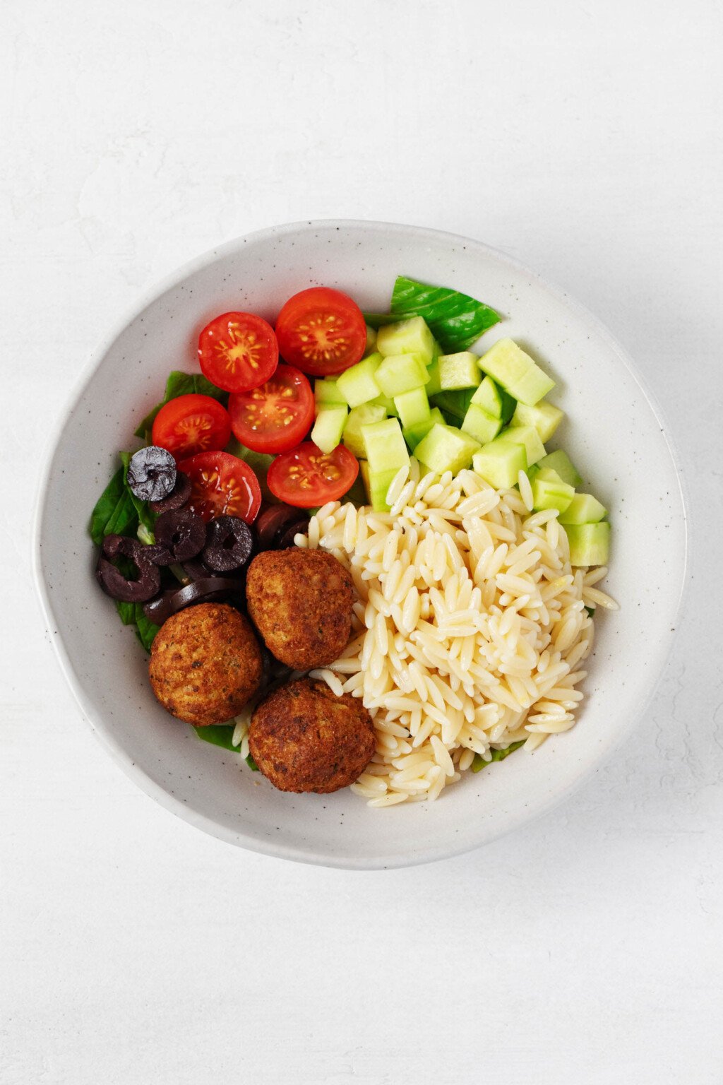 A vibrant vegan Greek bowl is being assembled in a white ceramic vessel. The bowl includes orzo, fresh vegetables, olives, and chickpea oat balls.
