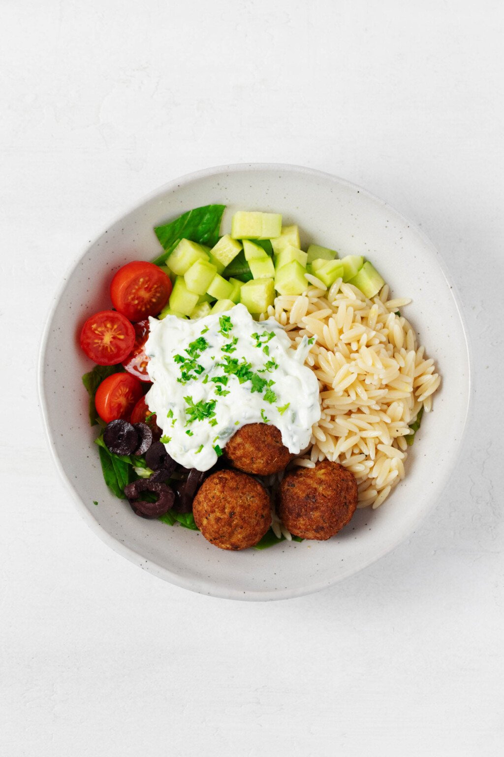 A vibrant vegan Greek bowl is being assembled in a white ceramic vessel. The bowl includes orzo, fresh vegetables, olives, and chickpea oat balls.