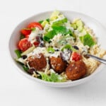 A white ceramic container holds a colorful vegan Greek bowl, including cooked orzo, tomatoes, and a cashew-based tzatziki.