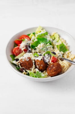 A white ceramic container holds a colorful vegan Greek bowl, including cooked orzo, tomatoes, and a cashew-based tzatziki.