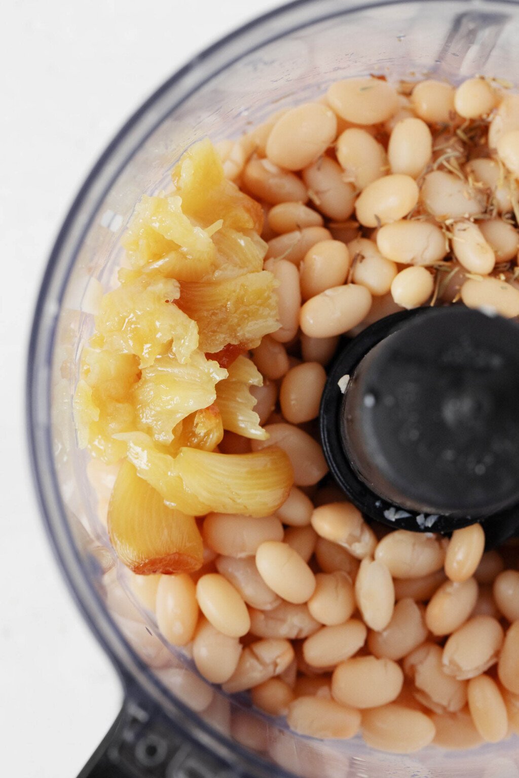The bowl of a food processor has been filled with white beans and roasted garlic.