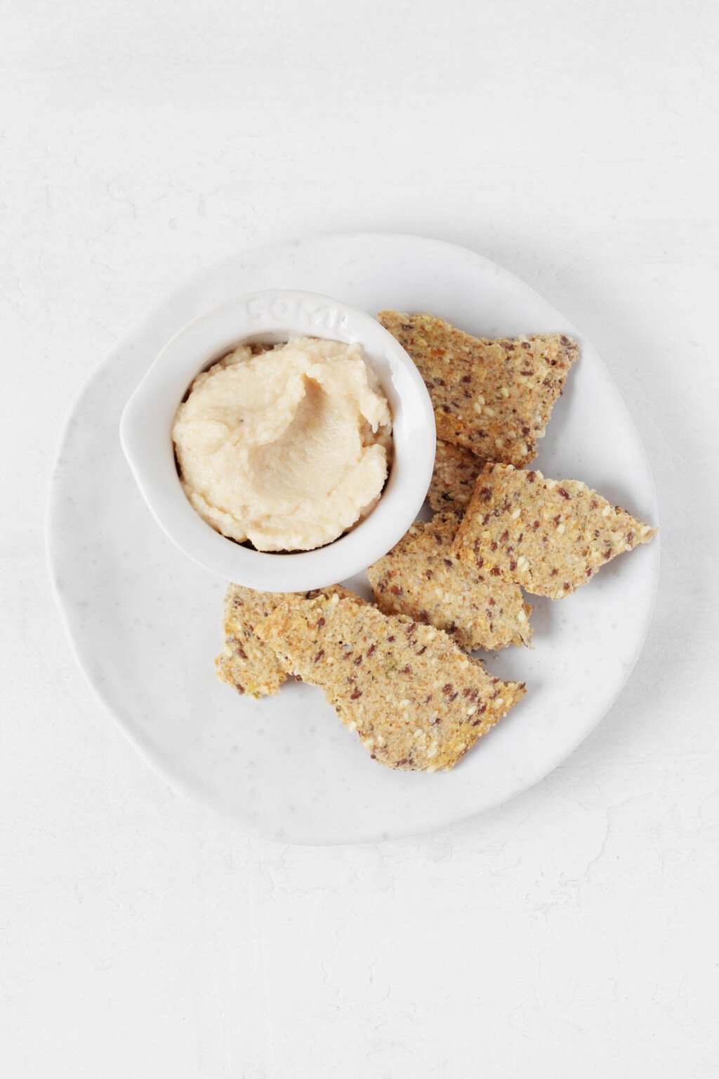 An overhead image of a small, pinch pot filled with a creamy white bean dip. The dip is served on a round white plate and accompanied by seeded crackers.